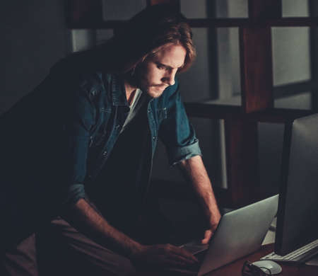 Handsome young businessman with shoulder-length blond hair and in casual wear is working with a computer at nightの写真素材