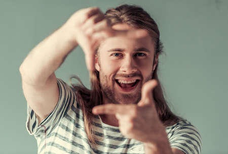 Stylish young man with shoulder-length blond hair is making frame at camera and smiling, on gray backgroundの写真素材