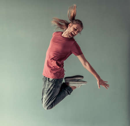 Stylish young man with shoulder-length blond hair and in casual clothes is screaming while jumping, on gray backgroundの写真素材