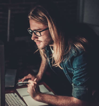 Handsome young businessman with shoulder-length blond hair and in casual wear and eyeglasses is making notes while working with a computer at nightの写真素材