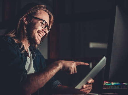 Handsome young businessman with shoulder-length blond hair is using a digital tablet and smiling while working with a computer at nightの写真素材