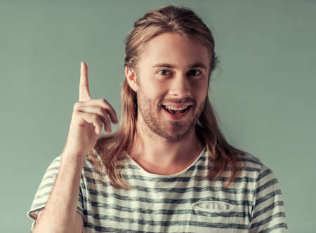 Stylish young man with shoulder-length blond hair is pointing upward, looking at camera and smiling, on gray backgroundの写真素材