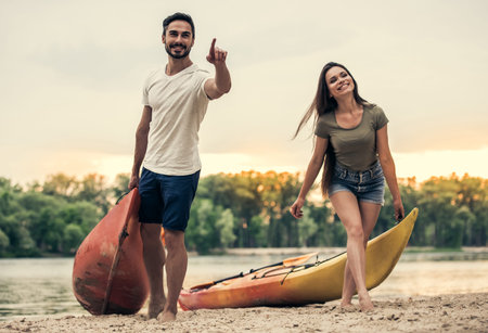 Happy young couple is talking and smiling while pulling kayaks on the beach after sailingの写真素材