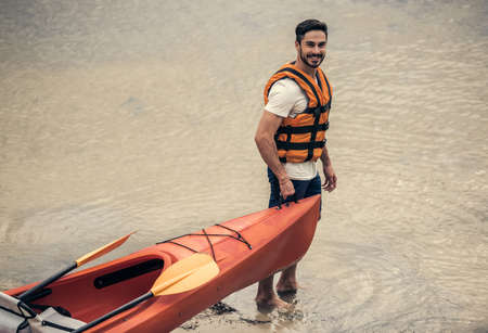 Handsome young man in sea vest is looking at camera and smiling while pulling a kayak to the river to sailの写真素材
