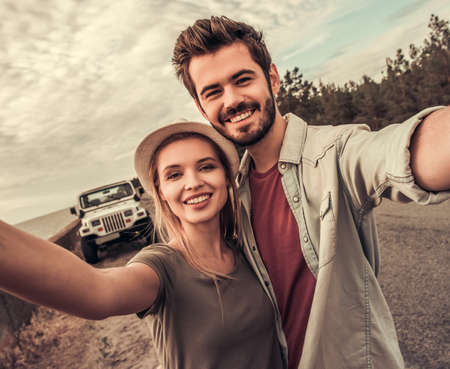 Beautiful couple is hugging, looking at camera and smiling while walking near the sea during their car tripの写真素材