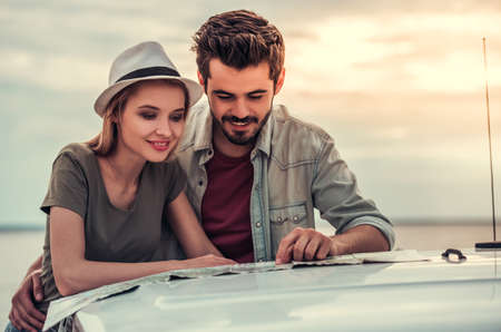 Beautiful couple is studying map and smiling while leaning on the hood of their car during tripの写真素材