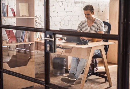 Handsome young businessman in casual clothes is using a laptop and smiling while working in office,...
