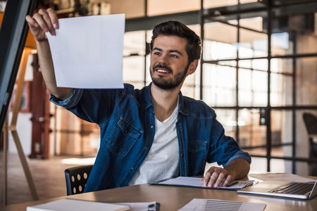 Attractive businessman in casual clothes is studying documents and smiling while working with a laptop in officeの写真素材
