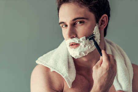 Handsome young man with foam on his face is shaving using a razor, on gray backgroundの写真素材