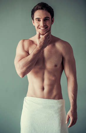 Handsome young man wrapped in bath towel and with bare torso is touching his chin, looking at camera and smiling, on gray backgroundの写真素材