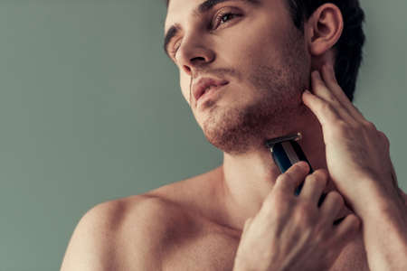 Handsome young man with bare torso is using an electric shaver, on gray backgroundの写真素材
