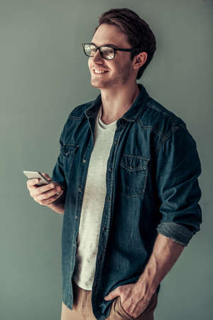 Handsome young man in casual clothes and eyeglasses is using a smart phone and smiling, on gray backgroundの写真素材