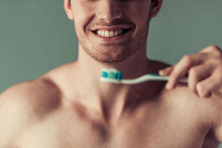 Cropped image of handsome young man with bare torso holding a toothbrush and smiling, on gray backgroundの写真素材
