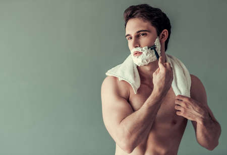 Handsome young man with foam on his face is shaving using a razor, on gray backgroundの写真素材