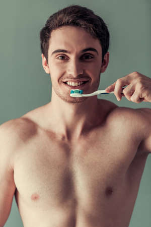 Handsome young man with bare torso is holding a toothbrush, looking at camera and smiling, on gray backgroundの写真素材