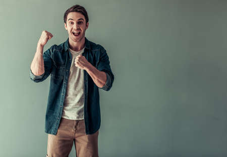 Handsome young man in casual clothes is raising hands in fists, looking at camera and shouting with happiness, on gray backgroundの写真素材