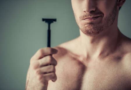 Cropped image of handsome young man with bare torso holding a razor, on gray backgroundの写真素材
