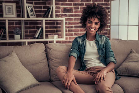 Beautiful Afro American girl is looking at camera and smiling while resting on couch at homeの写真素材