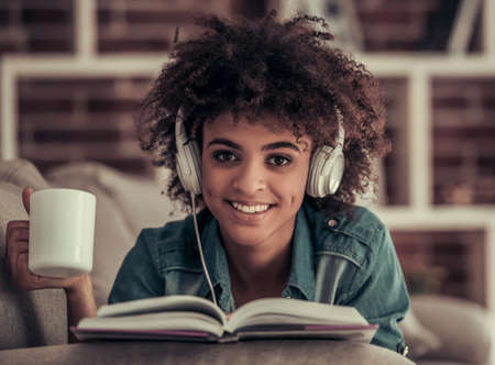 Beautiful Afro American girl in headphones is listening to music, reading a book, drinking coffee and smiling while resting at homeの写真素材