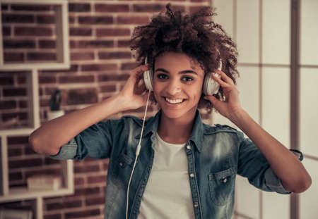 Beautiful Afro American girl in headphones is listening to music, looking at camera and smiling while spending time at homeの写真素材