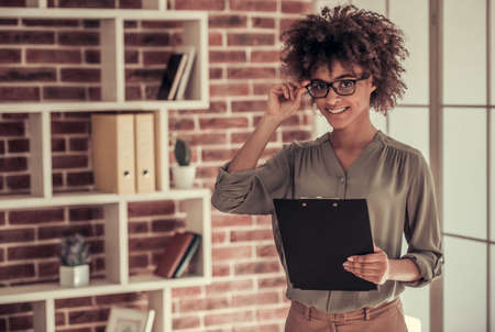 Beautiful Afro American business woman in smart casual wear and eyeglasses is looking at camera and smiling while standing with a folder in officeの写真素材