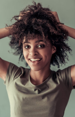 Portrait of beautiful Afro American girl playing with her curly dark hair, looking at camera and...