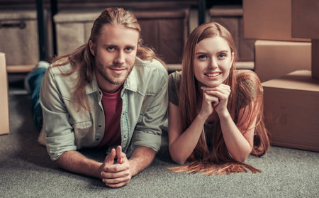 Beautiful young couple is looking at camera and smiling while lying on the floor among cardboard...