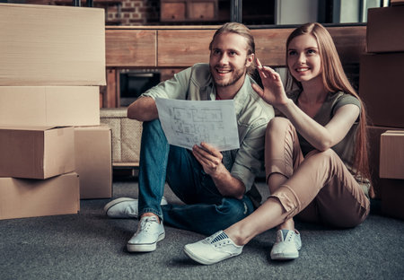 Beautiful young couple is holding an apartment plan and smiling while sitting on the floor among...