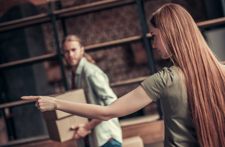 Young couple is drifting apart after breakup, girl is pointing to the entrance, man is carrying his stuff in cardboard boxesの写真素材