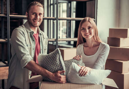 Beautiful young couple is packing their stuff into cardboard boxes and smiling while moving into new apartmentの写真素材