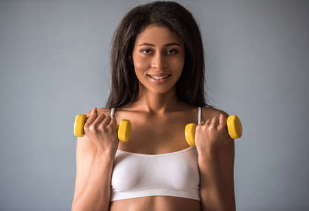 Beautiful Afro American sports girl is holding dumbbells, looking at camera and smiling, on gray backgroundの写真素材