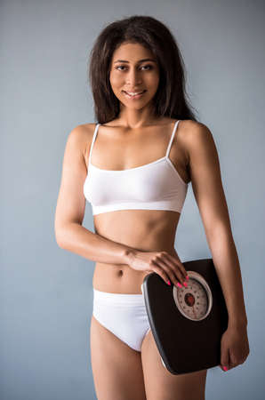 Beautiful Afro American sports girl is holding a weigh scales, looking at camera and smiling, on gray backgroundの写真素材