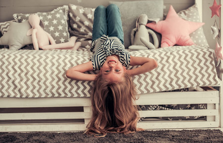 Pretty little girl is looking at camera and smiling while lying on her back on bed at homeの写真素材