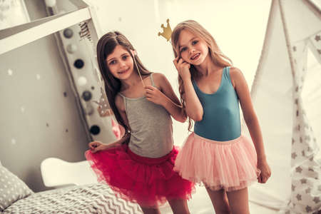 Two happy little girls in crowns are holding party props, looking at camera and smiling while playing in children's room at homeの写真素材