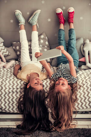 Two cute little girls are using a digital tablet and smiling while lying on their backs on bed in children's roomの写真素材