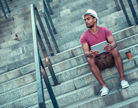 Handsome Afro American man is using a digital tablet while sitting on stairs during city walk