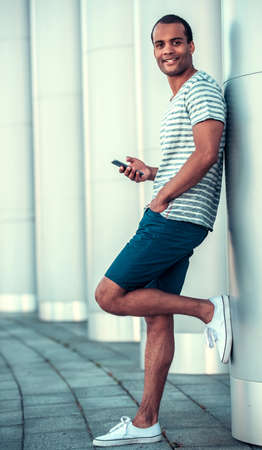 Full length portrait of handsome Afro American man using a smart phone, looking at camera and smiling while walking in cityの写真素材
