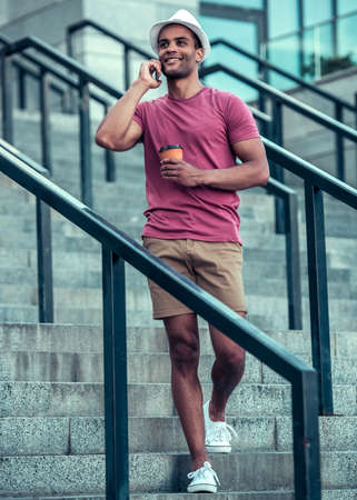Handsome Afro American man is talking on the mobile phone, holding a cup of coffee and smiling while walking in cityの写真素材