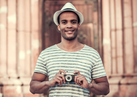 Handsome Afro American man is holding a photo camera and smiling while walking in cityの写真素材