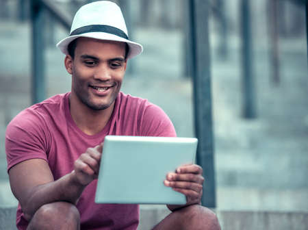 Handsome Afro American man is using a digital tablet and smiling while sitting on stairs during city walkの写真素材