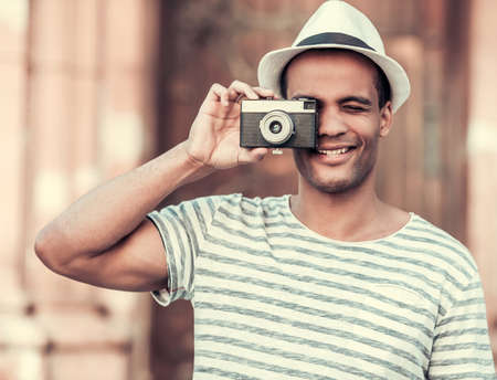 Handsome Afro American man is holding a photo camera and smiling while walking in cityの写真素材