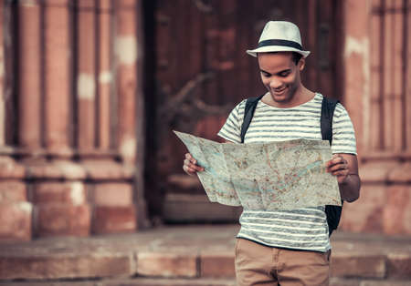 Handsome Afro American man with backpack is examining a map and smiling while walking in new cityの写真素材