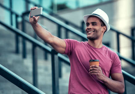 Handsome Afro American man is doing selfie using a smart phone, holding a cup of coffee and smiling while walking in cityの写真素材