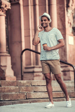 Full length portrait of handsome Afro American man talking on the mobile phone, holding a cup of coffee and smiling while walking in cityの写真素材