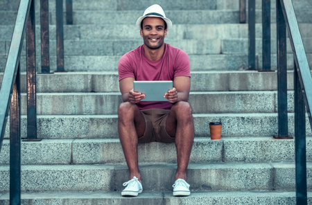 Handsome Afro American man is using a digital tablet, looking at camera and smiling while sitting on...