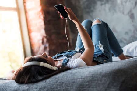Beautiful Afro American girl in denim overall and headphones is listening to music using a smart phone while lying on bed at homeの写真素材