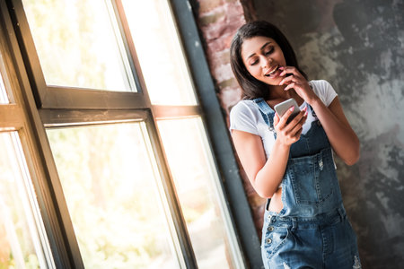 Beautiful Afro American girl in denim overall is using a smart phone and smiling while standing in...