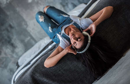 Beautiful Afro American girl in denim overall and headphones is listening to music and smiling while lying on bed at homeの写真素材