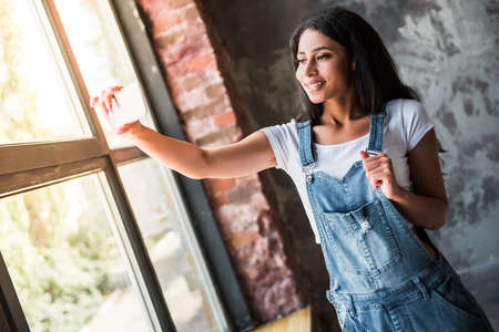 Beautiful Afro American girl in denim overall is doing selfie using a smart phone and smiling while standing in room at homeの写真素材