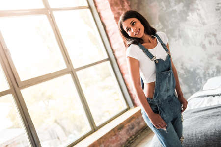 Beautiful Afro American girl in denim overall is smiling while standing in room at homeの写真素材